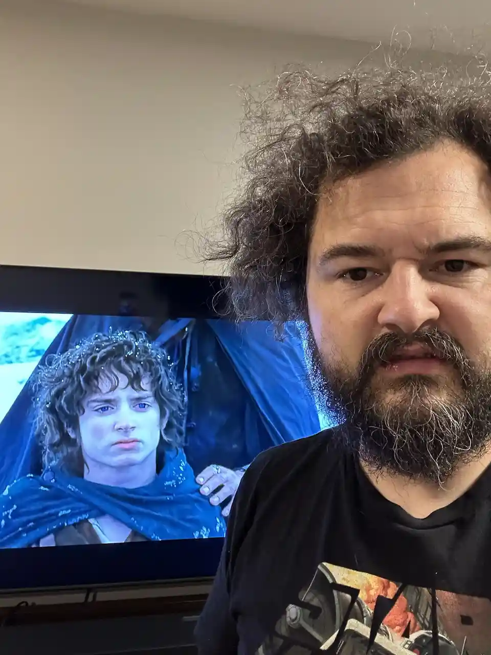 A man with a beard and curly hair stares intensely at the camera, positioned in front of a TV screen displaying a scene from…