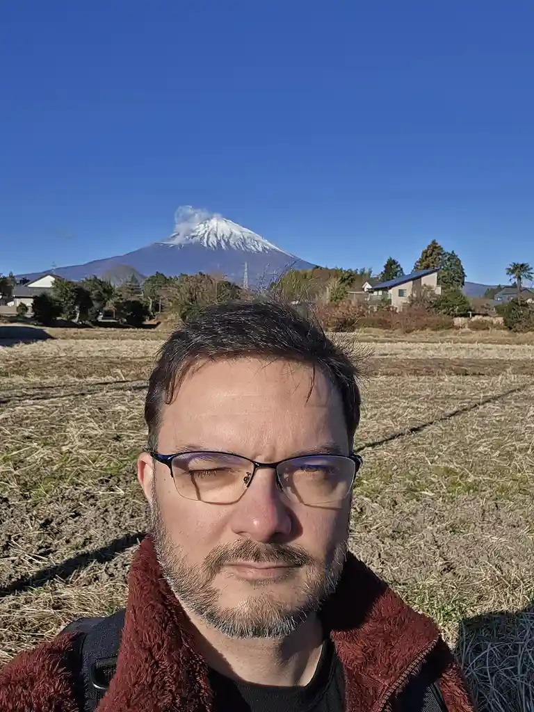 A man with glasses and a beard, wearing a red fleece jacket, takes a selfie with his eyes squeezed shut and a slight…