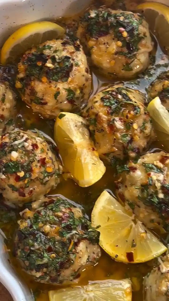 A close-up photo of several round, golden-brown meatballs in a white baking dish, covered in a glossy sauce and garnished…