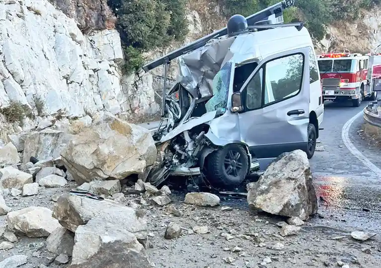 A silver van is severely damaged after crashing into a rocky embankment on a winding road, with large boulders scattered…