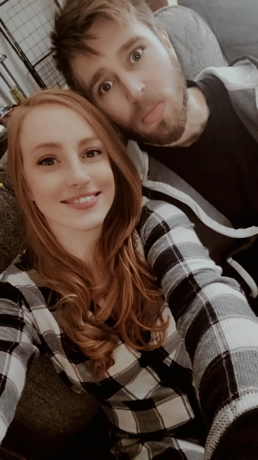 A selfie of a smiling woman with red hair and a nose ring wearing a plaid shirt, taking a photo with a man behind her who…