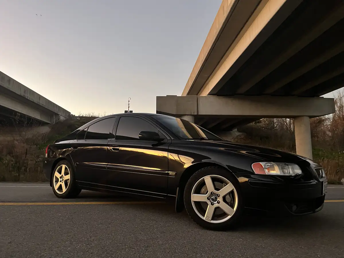 A black Volvo S60 sedan is parked on a road under concrete overpasses during sunset, its glossy paint reflecting the golden…