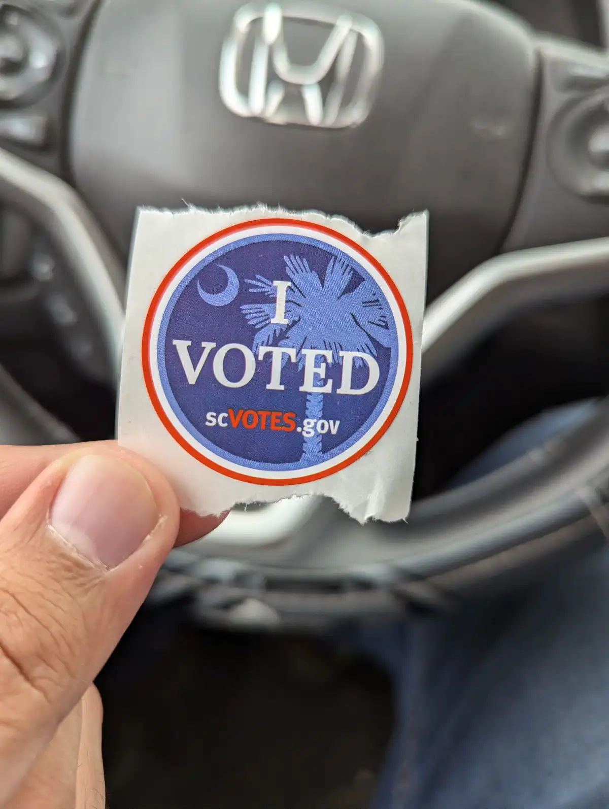 A hand holds up a torn 'I VOTED' sticker from South Carolina, featuring a palm tree and crescent moon, in front of a car's…