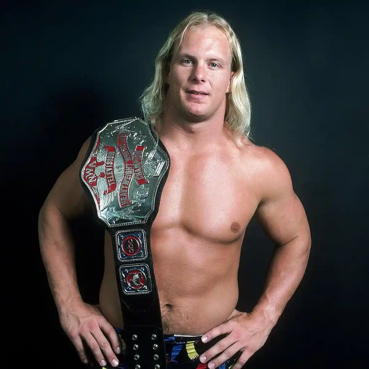 a shirtless, muscular man with long blonde hair poses confidently with his hands on his hips, wearing a championship belt…