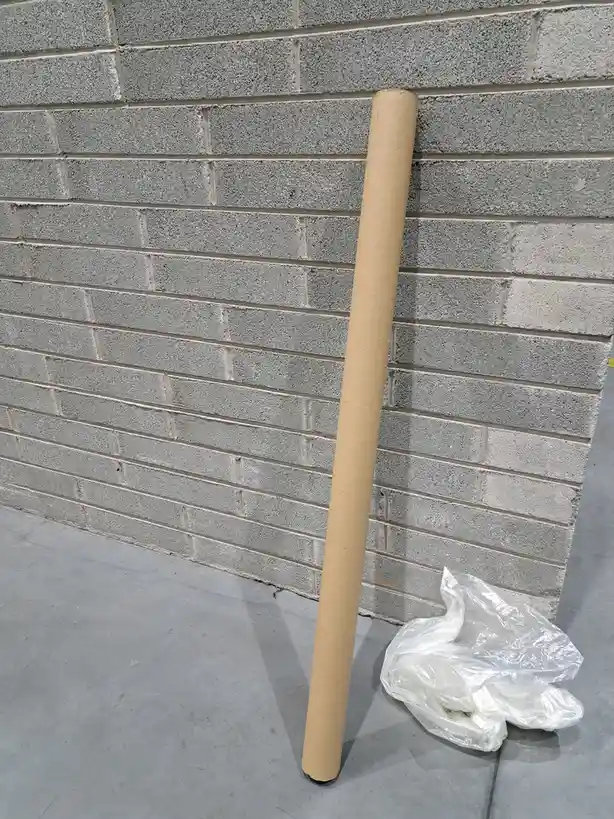 A tall, brown cardboard tube leans against a gray brick wall on a concrete floor, with a crumpled white plastic bag nearby…