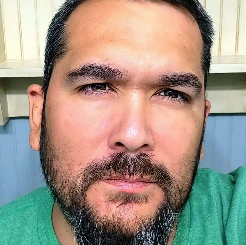 A close-up selfie of a man with a beard and mustache, wearing a green shirt, looking directly at the camera with a stern…