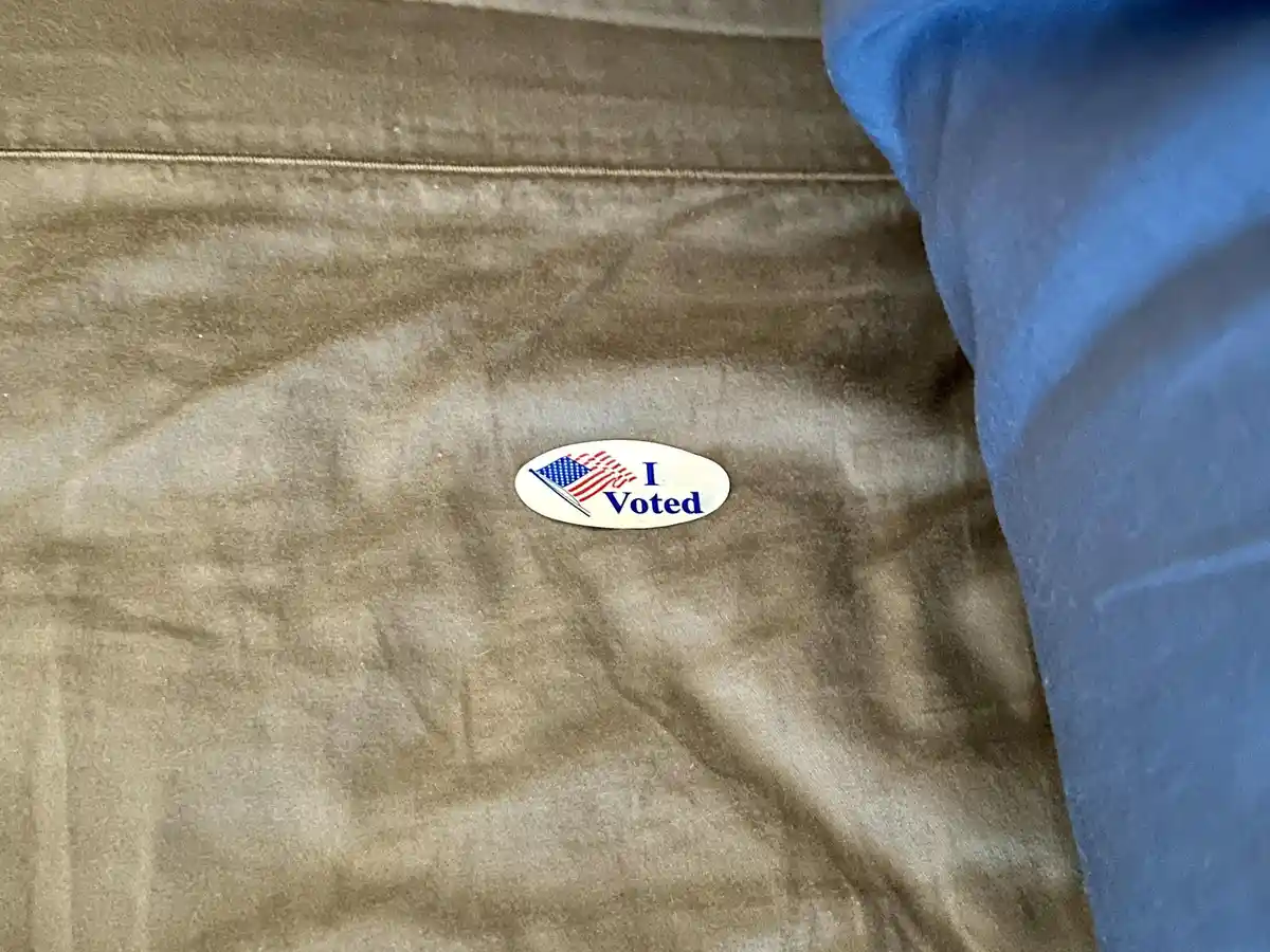 A close-up photo shows a small oval 'I Voted' sticker with an American flag design resting on a textured brown fabric…