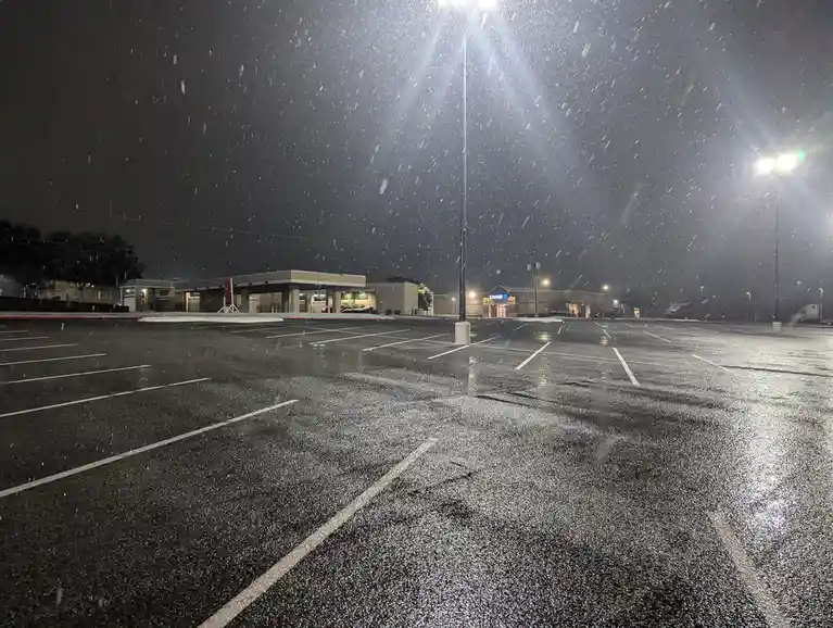 A nighttime photo of an empty parking lot with snow falling heavily, illuminated by bright overhead lights. In the…