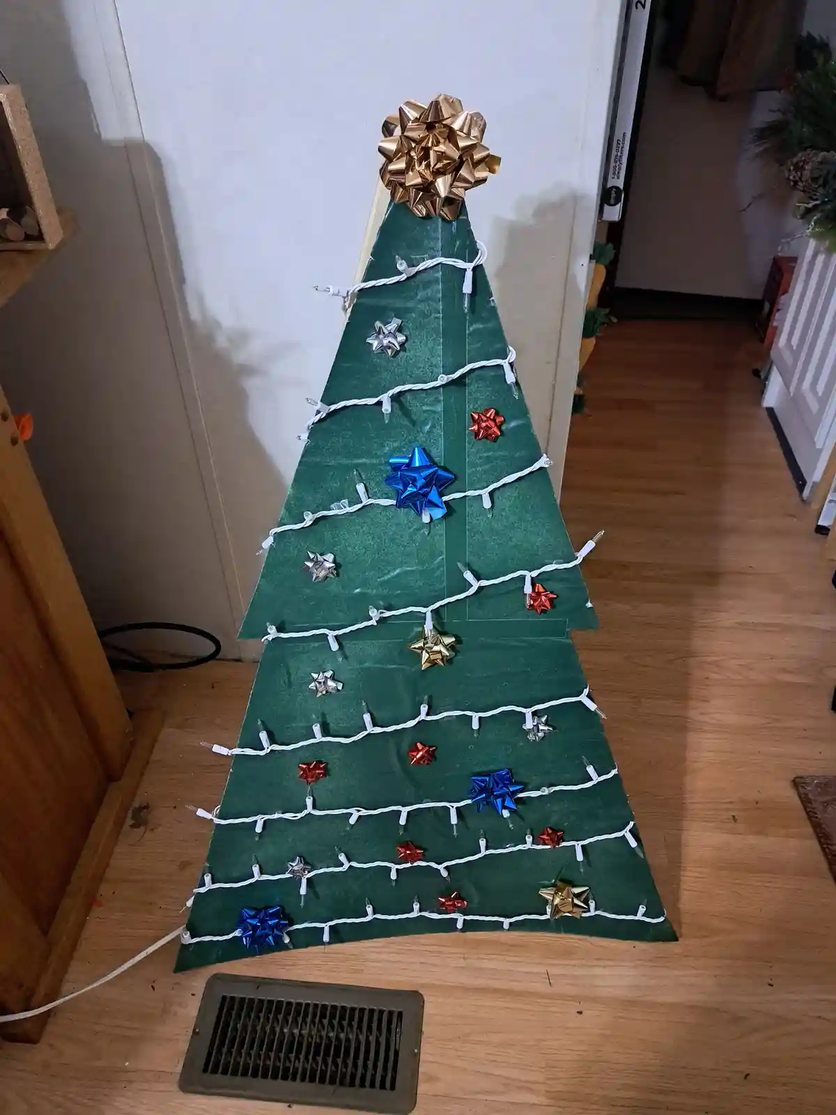 A Christmas tree made from green duct tape, standing upright on a wooden floor, decorated with white string lights and shiny…
