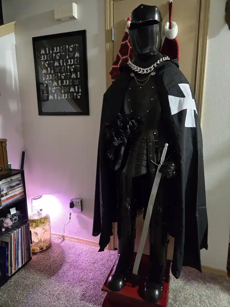 A full suit of black knight armor stands on a wooden pedestal in a room, wearing a black cape with a white cross on the…