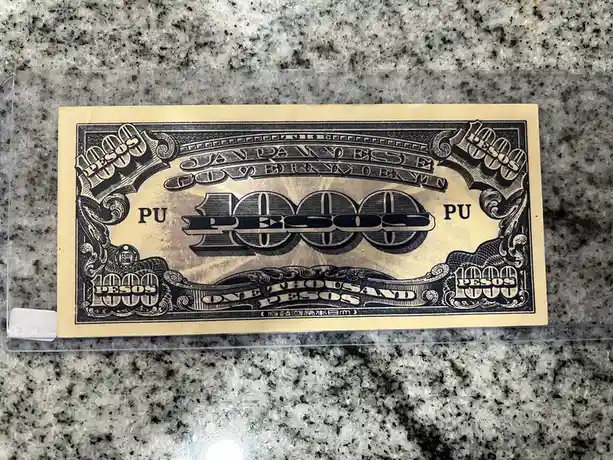 A close-up photo shows a yellowish paper bill with ornate black ink designs, resting on a speckled granite countertop inside…