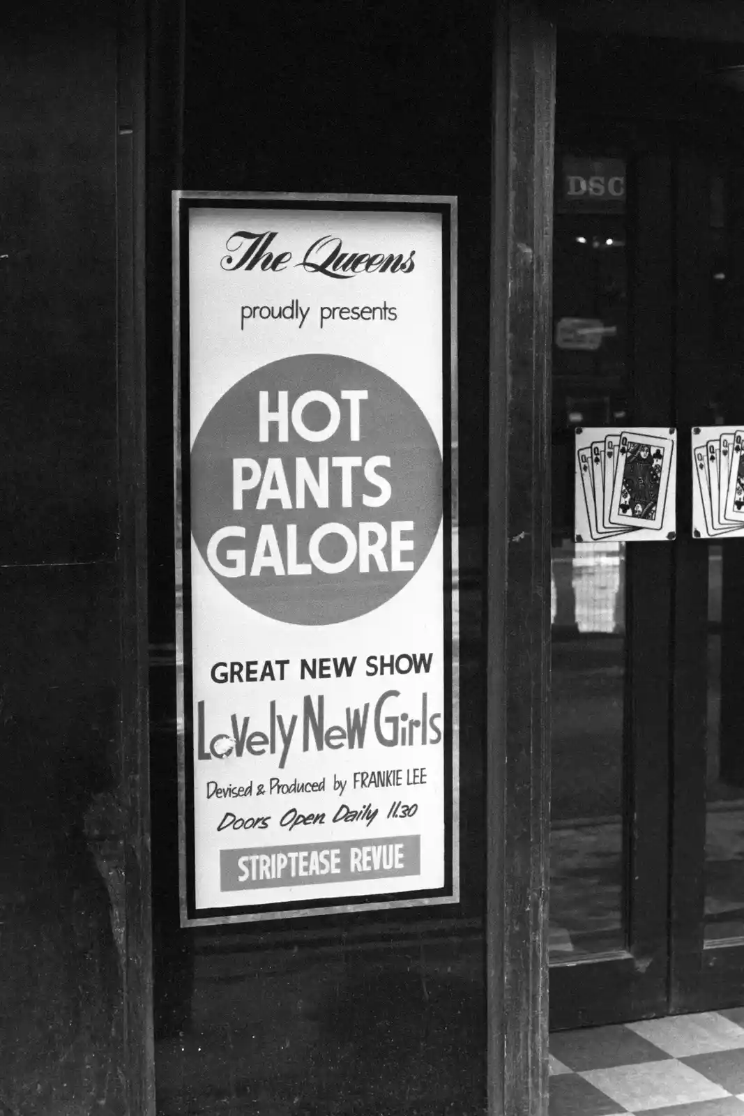 A black-and-white photo shows a vintage poster on a glass door advertising 'The Queens' strip club show 'Hot Pants Galore'…