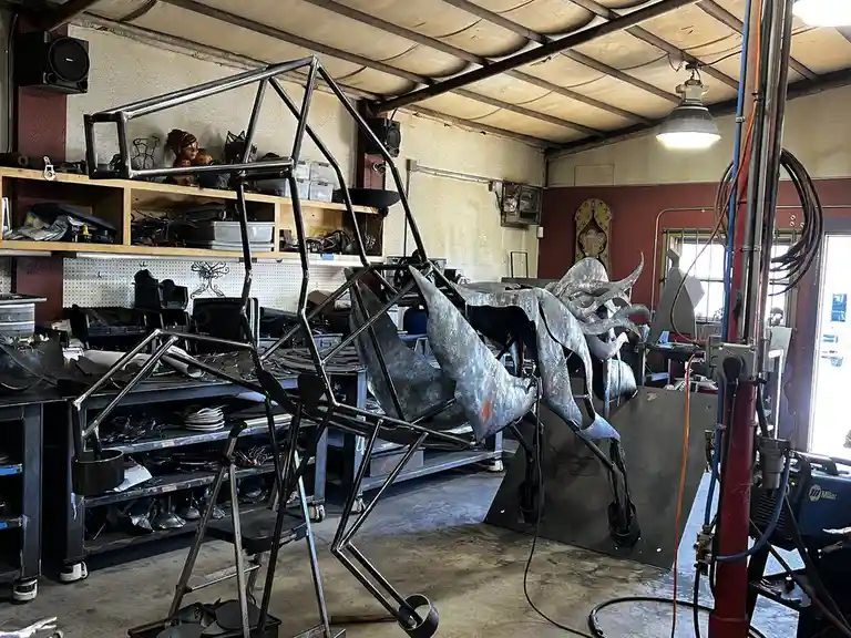 A cluttered metal workshop filled with welding equipment, shelves of tools, and large metal sculptures in various stages of…