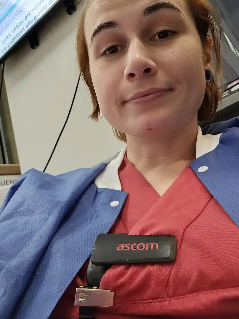 A close-up selfie of a woman in red and blue scrubs with a name badge that reads 'ascom' clipped to her chest. She has a…