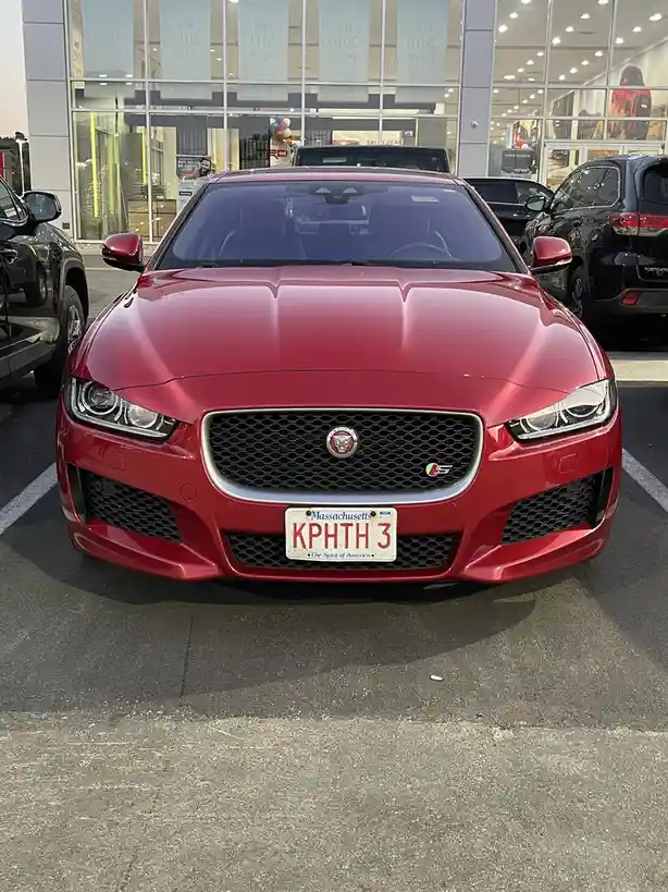 A red Jaguar car is parked in front of a dealership with a Massachusetts license plate reading 'KPHTH 3'. The car's front…