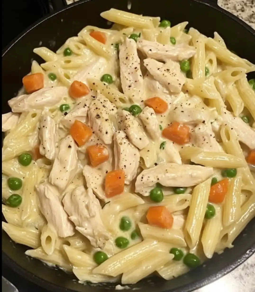 a close-up photo of creamy penne pasta mixed with shredded chicken, diced carrots, and green peas in a black skillet…