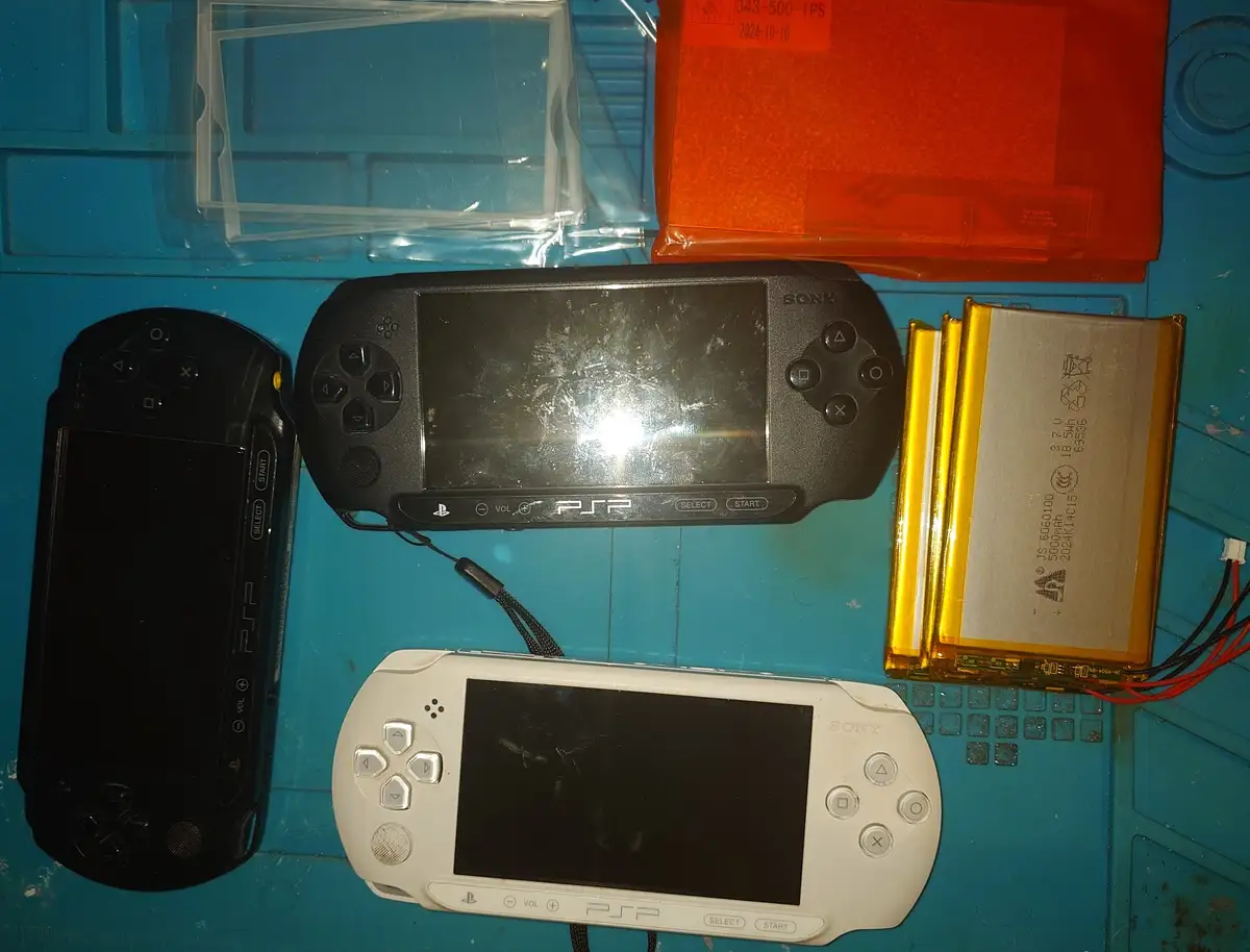 Three Sony PSP handheld gaming consoles (black, black, white) are laid out on a blue work surface alongside replacement…