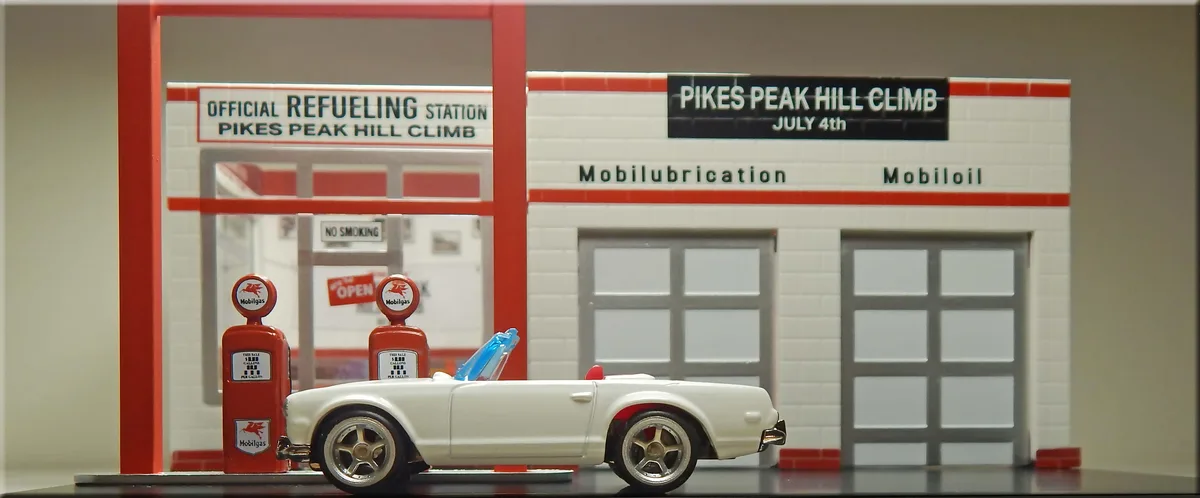A miniature model of a white convertible sports car is parked at a vintage Mobilgas gas station, which is labeled as the…