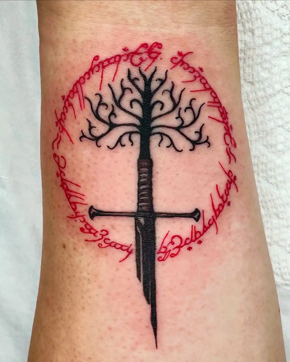 A fresh tattoo on skin shows a black tree with intricate branches, its trunk replaced by a sword hilt and blade, encircled…