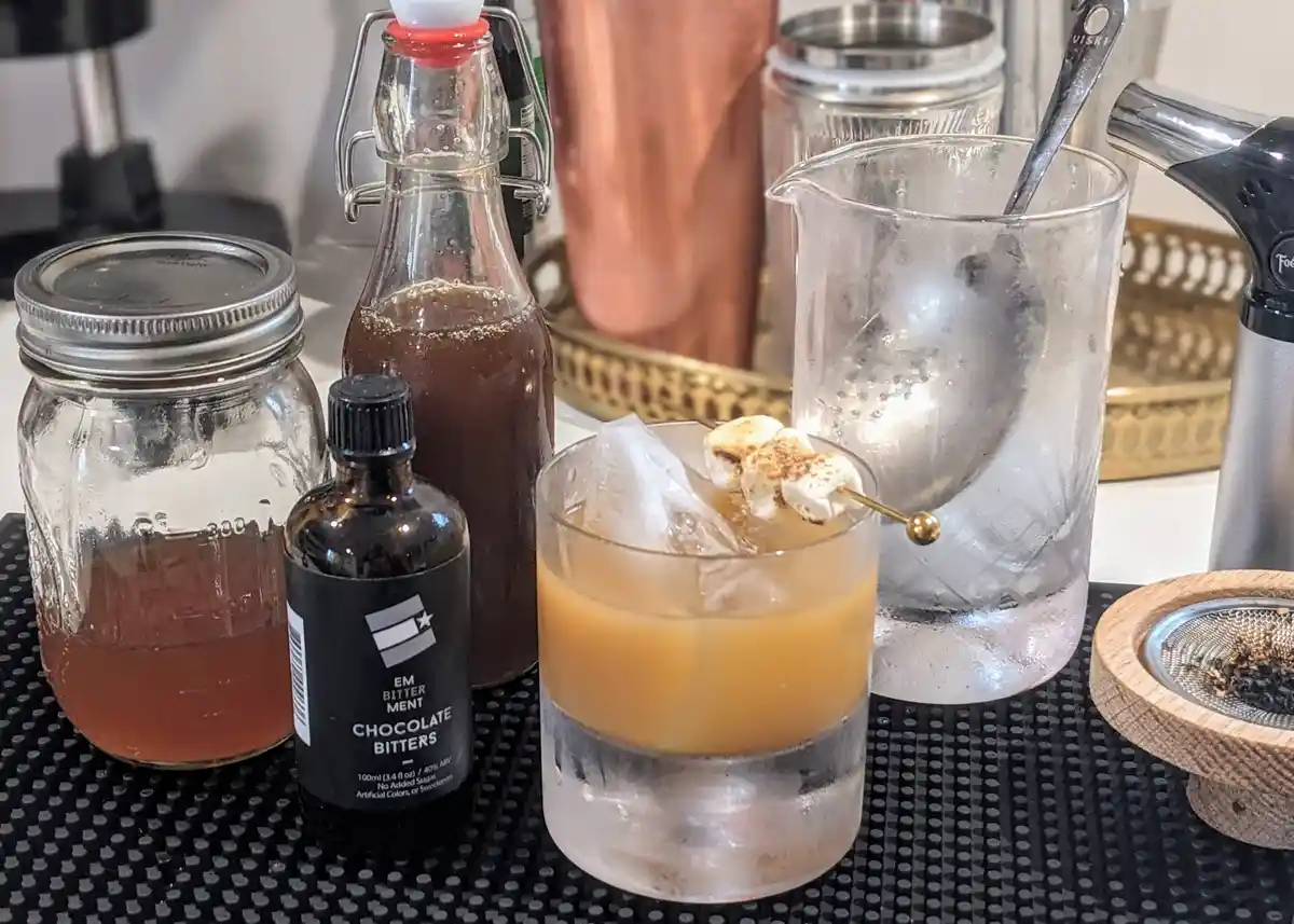 A cocktail in a rocks glass with ice and toasted marshmallows on a skewer sits on a bar mat, surrounded by ingredients…