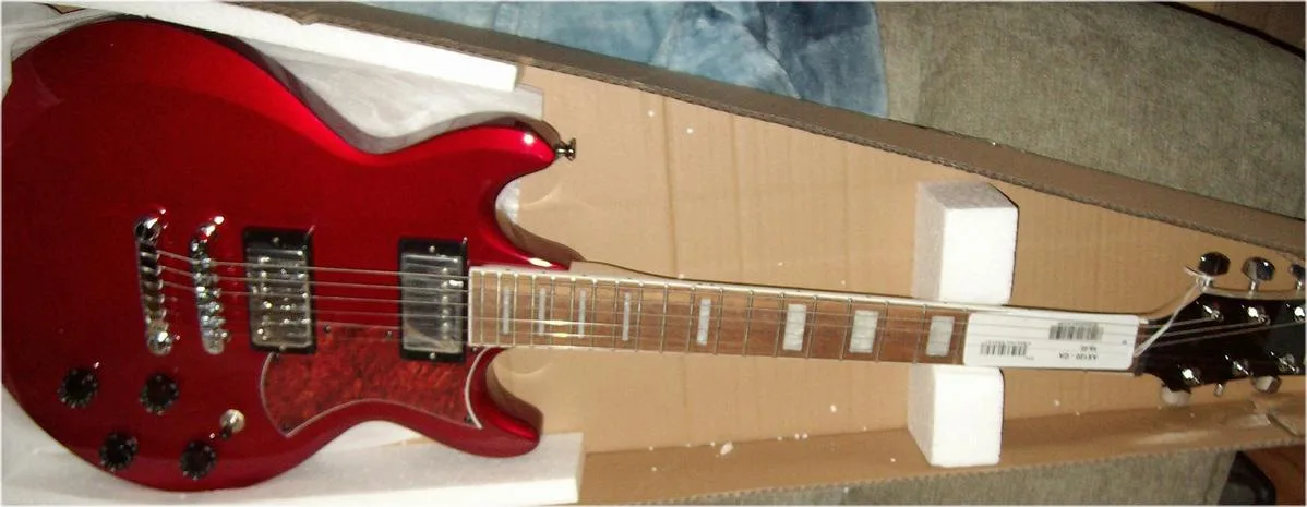 A red electric guitar is shown resting inside its cardboard packaging, propped up by white foam blocks. The guitar has a…