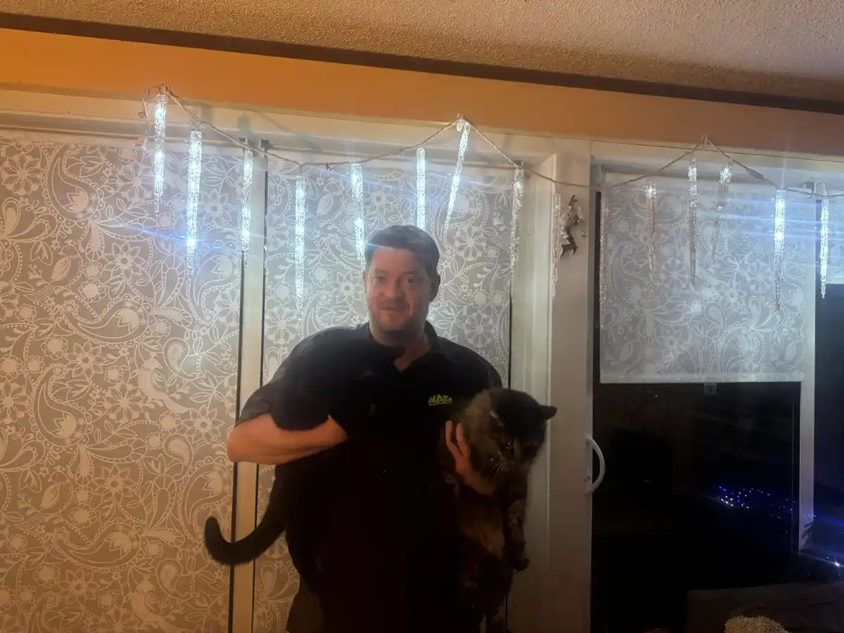 A man in a black shirt stands indoors holding two cats, one black and one tabby, in front of patterned curtains with icicle…