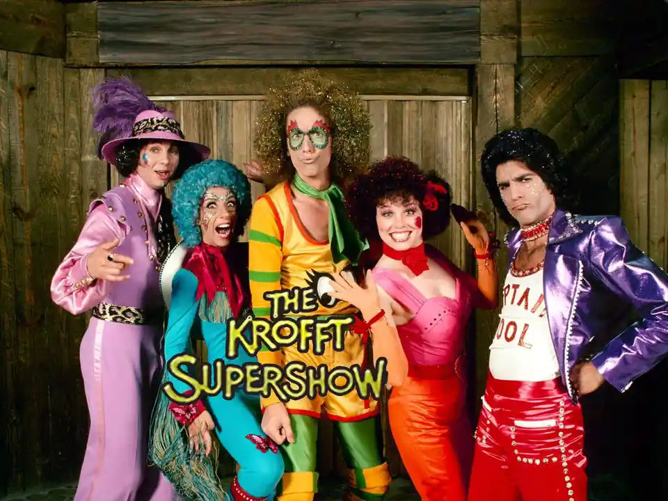 Five people in wildly colorful, 1970s-style costumes and heavy makeup pose in front of a wooden wall. The person on the far…