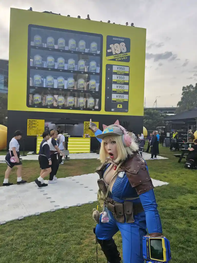 A person in a blue and brown Fallout-style cosplay with pink cat ears and headphones points at a giant yellow vending…
