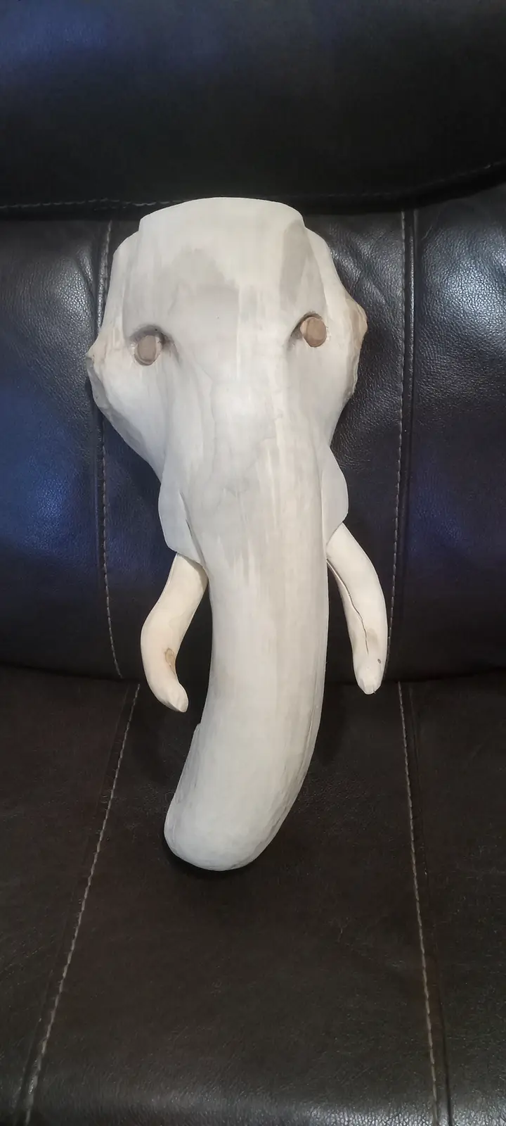 A white, hand-carved driftwood sculpture shaped like an elephant's head with two small round eyes and two curved tusks rests…