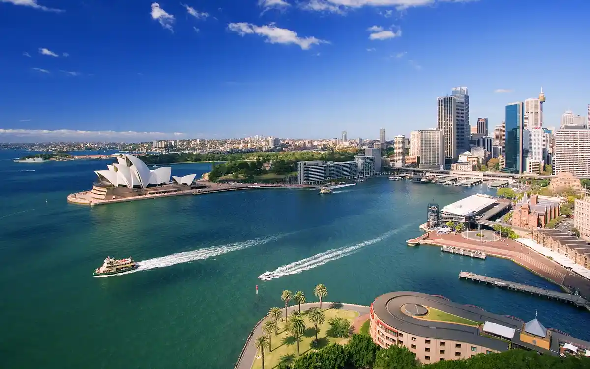 A scenic aerial photograph of Sydney Harbour, featuring the iconic white sail-like roof of the Sydney Opera House on the…