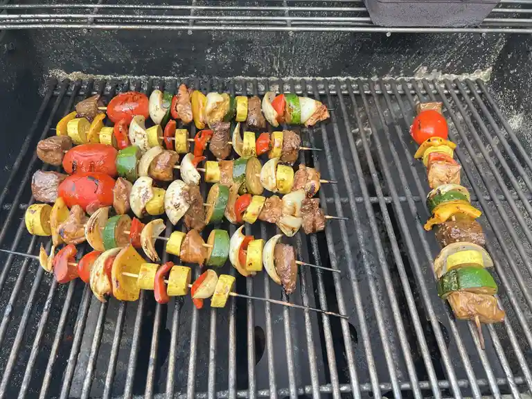 A close-up photo shows numerous colorful vegetable and meat skewers arranged on a metal grill grate, with some showing char…