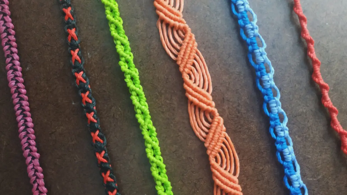 A close-up photo shows six colorful handmade friendship bracelets laid out side-by-side on a dark brown textured surface…