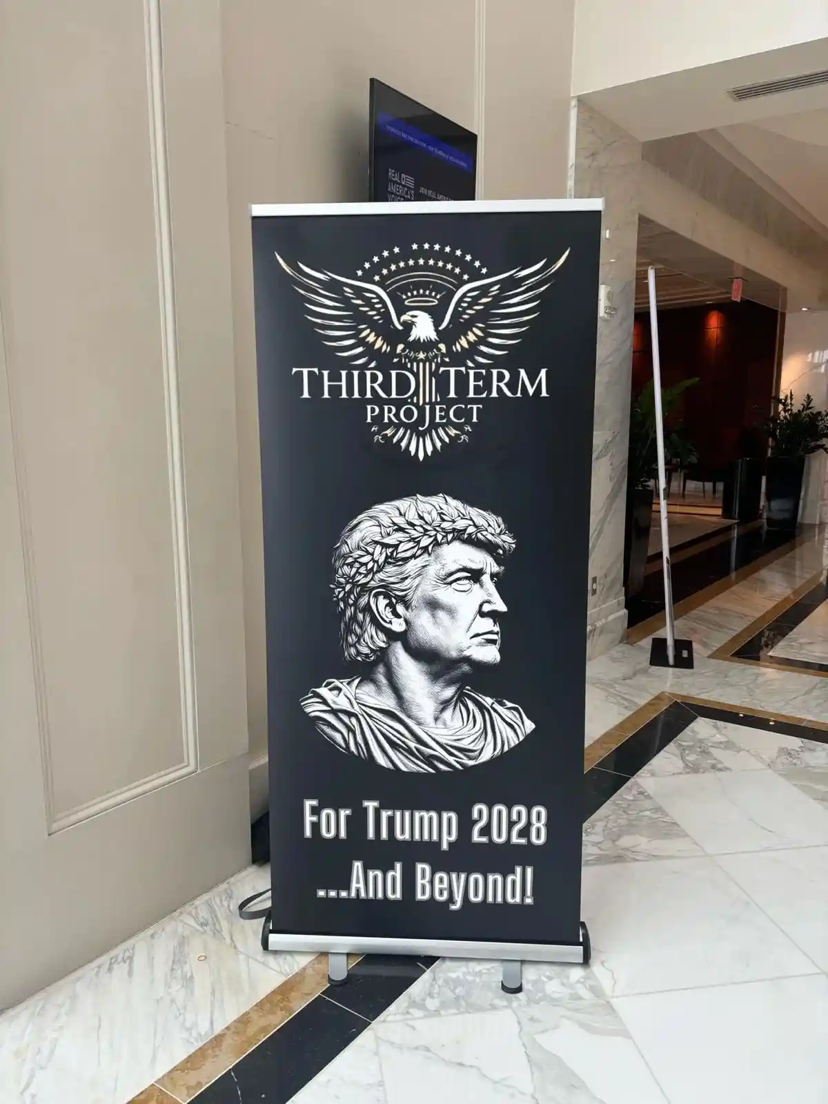 A black banner stand in a marble-floored lobby displays a logo of an eagle with stars above the text 'THIRD TERM PROJECT'…