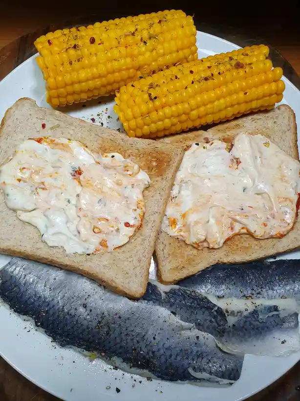 A white plate holds two slices of toasted bread topped with creamy orange sauce, two ears of seasoned corn on the cob, and…