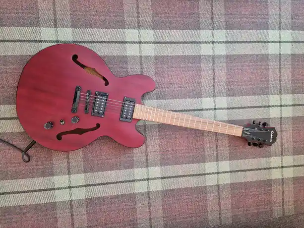 A red electric guitar with a cutaway body and two humbucker pickups lies on a plaid blanket with pink, gray, and beige…