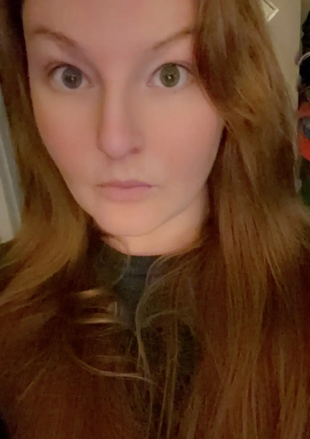 A close-up selfie of a woman with long brown hair and wide, startled green eyes looking directly at the camera. She has a…