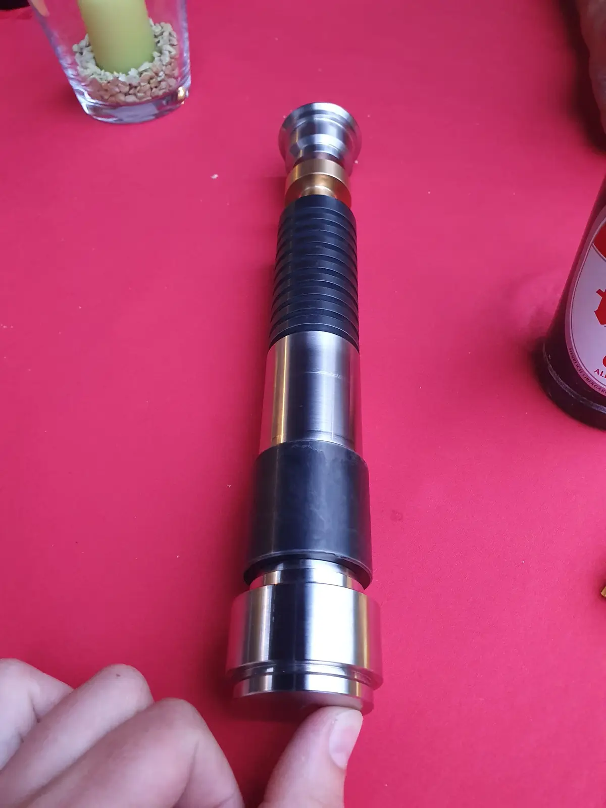 A close-up photo shows a person's hand holding a metallic, segmented lightsaber hilt with black grip sections against a red…