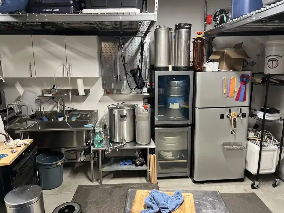 A homebrewing setup in a garage features stainless steel brewing kettles, kegs, and refrigerators with beer taps. Shelves…
