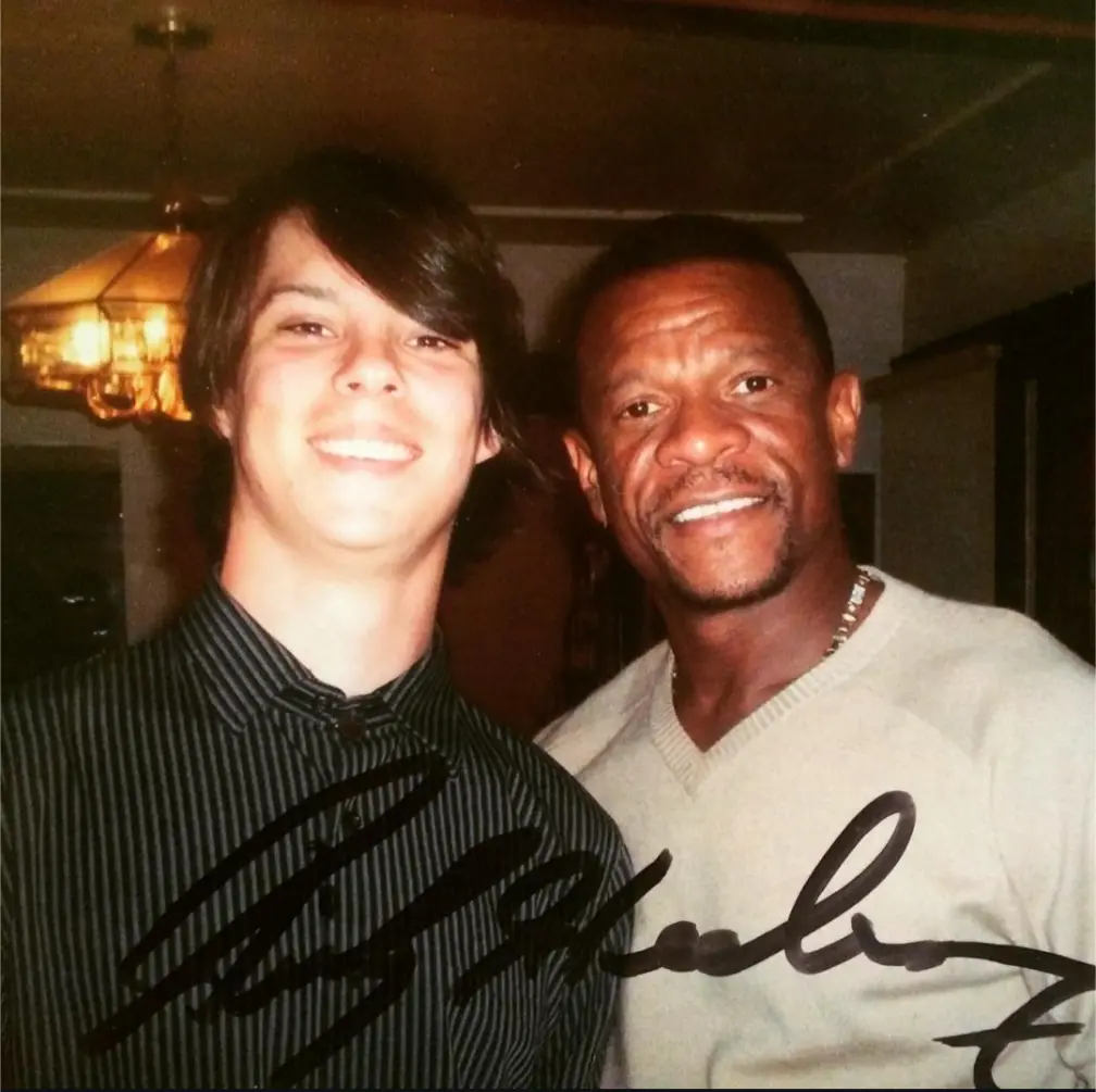 two men are smiling for a photo in a warmly lit indoor setting, one younger with dark hair in a striped shirt and one older…