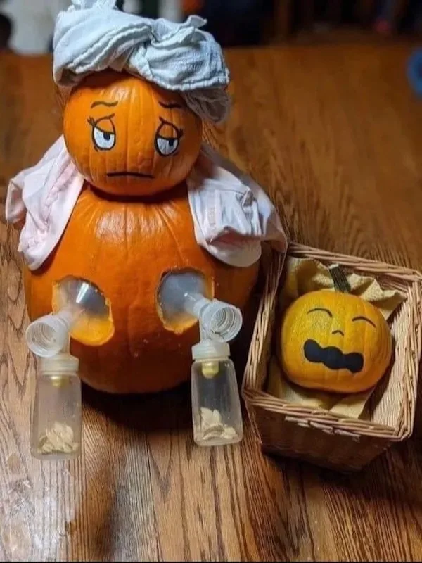 A carved pumpkin with a sad face, wearing a cloth head wrap and a white shirt, has two breast pumps attached to holes in its…