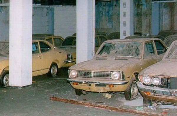 A dusty, abandoned car in a neglected parking garage with other similarly neglected vehicles. The car in the foreground has…