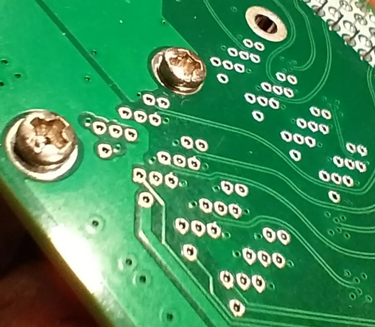 A close-up photo of a green computer motherboard showing two screws with damaged heads and numerous empty solder pads…