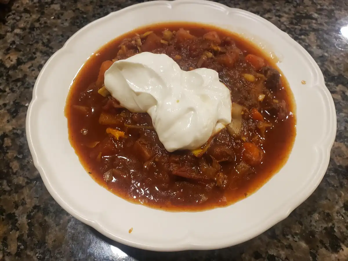 A white scalloped bowl filled with a thick, reddish-brown stew containing visible chunks of meat and vegetables, topped with…