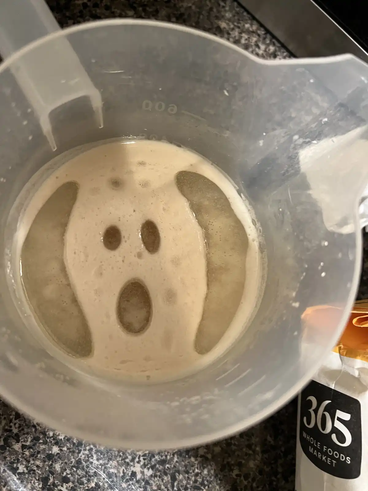 A close-up photo shows a clear plastic measuring cup containing a light-brown foamy liquid, where the foam has naturally…