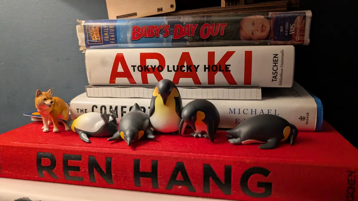 A stack of books and a VHS tape are arranged on a shelf, with several small penguin figurines and one small dog figurine…
