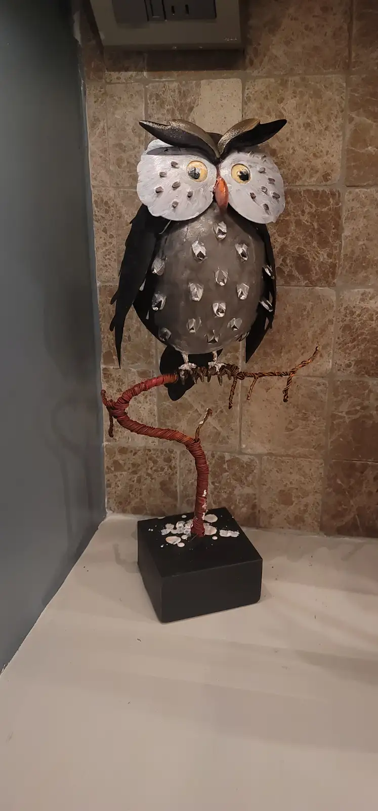 A metal owl sculpture with large white eyes and a gray body adorned with silver gems sits perched on a twisted wire branch…