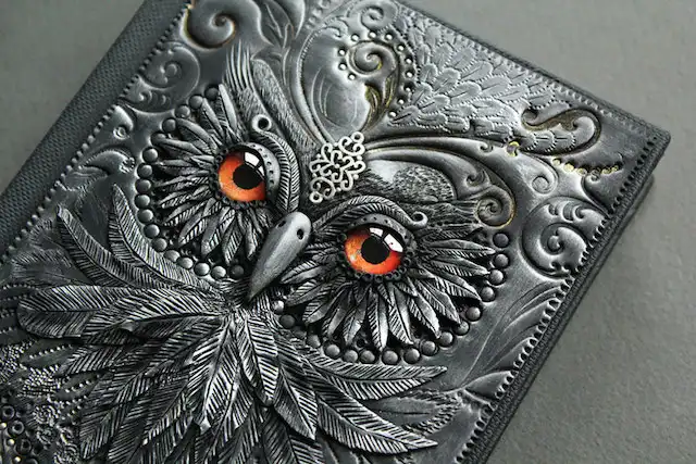A close-up of an intricately embossed leather wallet or journal cover featuring a highly detailed owl design. The owl is…