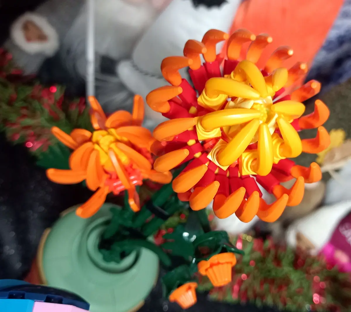 A close-up photo shows two bright, colorful flowers made entirely of LEGO bricks, with orange, red, and yellow petals and…