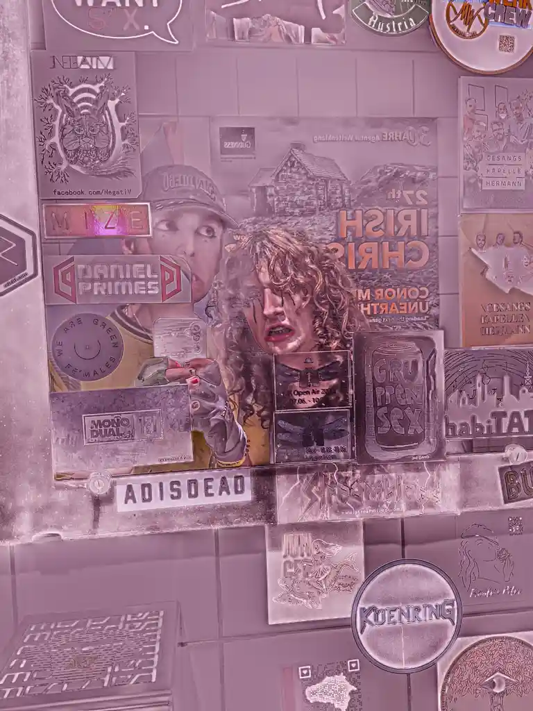 A wall covered in various metal-looking stickers and posters, including one of a person with curly hair and dramatic makeup…