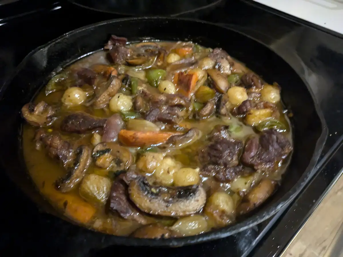 A close-up photo shows a black cast-iron skillet on a stovetop, filled with a stew containing chunks of beef, sliced…